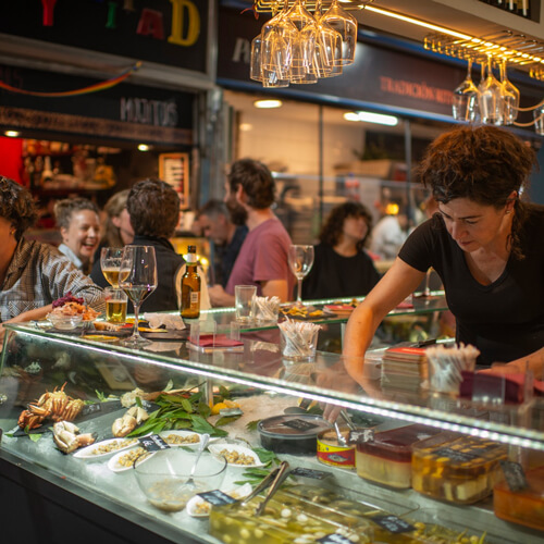 Mujer atendiendo barra de bar con clientes hablando entre sí