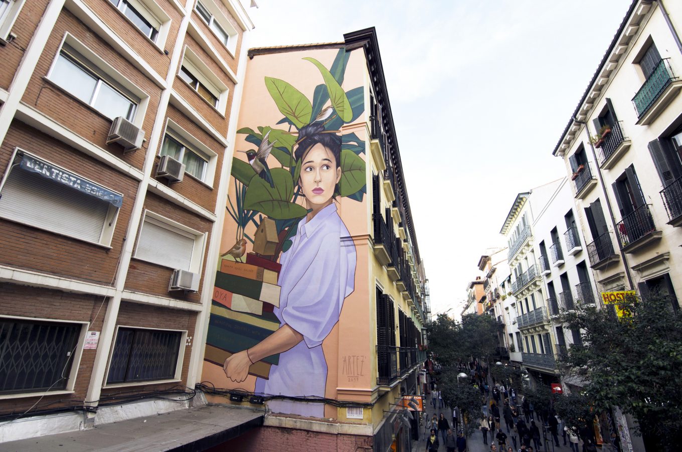 Mural mujer acarreando libros y planta en fachada de edificio